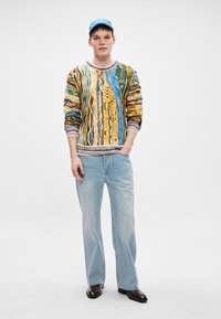 Monivärinen neulepaita, jossa on monimutkaisia kuvioita, yhdistettynä vaaleansinihin farkkuihin ja mustiin teräväkärkisiin kenkiin. Vaaleansininen hattu antaa kontrastia.