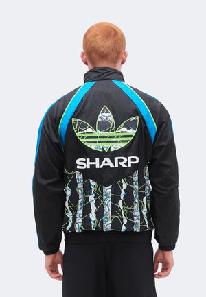 Osoba s kratkom crvenom kosom koja nosi crnu jaknu s plavim prugama i velikim zelenim logom "SHARP" i uzorkom na leđima, okrenuta leđima.