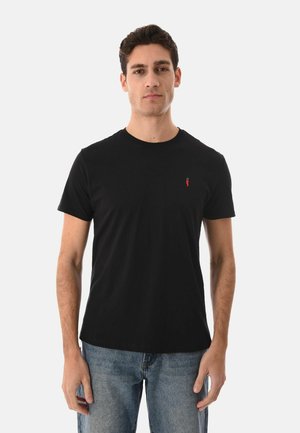 Uomo che indossa una t-shirt nera a maniche corte con un piccolo logo rosso sul petto e jeans blu, in piedi contro uno sfondo bianco.
