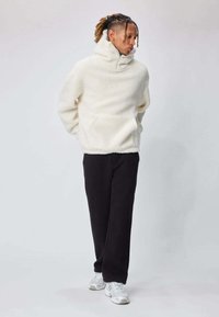 Pull-over en polaire blanche avec col montant, poche avant et poignets côtelés, assorti à un pantalon noir coupe décontractée et des baskets blanches.