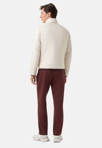 Helle beige isolierte Jacke mit Stehkragen, gerippten Bündchen und schwarzem Besatz, kombiniert mit bordeauxroten Jogginghosen und weißen Sneakers.