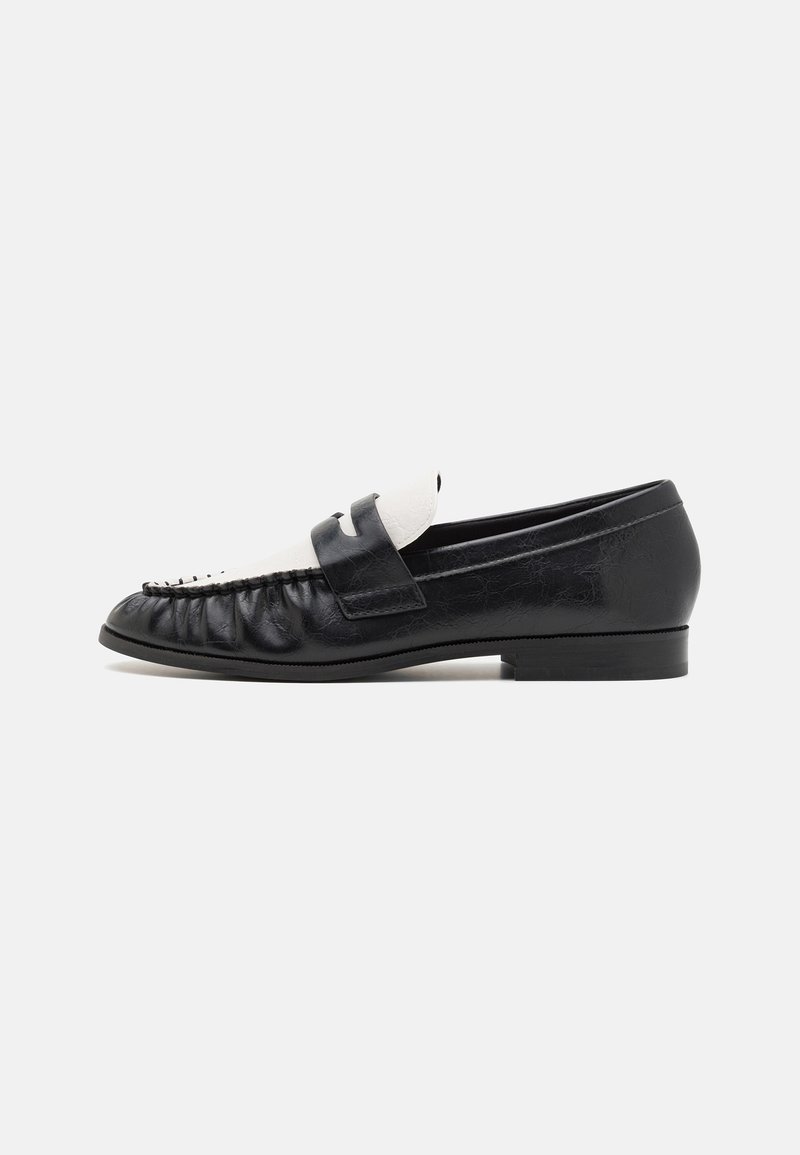 Mocassin en cuir noir avec incrustation en cuir blanc et bride à penny, semelle plate, et coutures de style mocassin sur un fond gris clair.