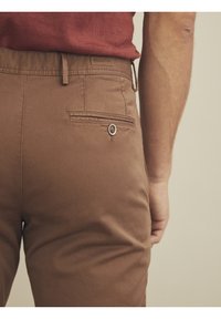 Celana cokelat dengan tekstur halus dan saku belakang yang dilengkapi dengan penutup kancing. Dipadukan dengan kemeja merah lengan pendek.
