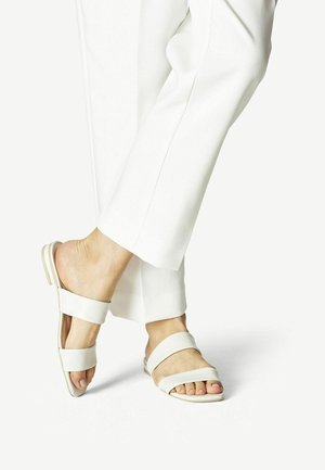 Sandales blanches à semelles plates avec deux sangles réglables et un design ouvert sur l'avant. Portées avec un pantalon blanc ajusté devant un fond neutre.