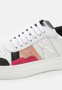 Baskets blanches avec des empiècements en suède noir, rose et beige, lacets blancs, emblème texturé d'abeille et texte "LAISSE-MOI VOLER" sur la semelle latérale.