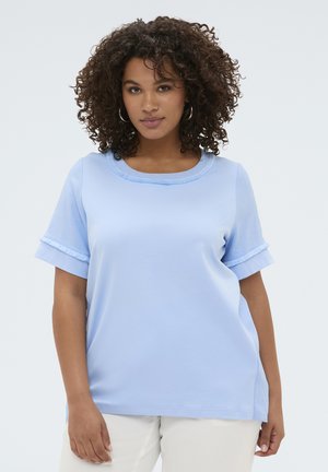 Kvinde med krøllet hår iført lyseblå top med korte ærmer og flæsedetaljer på kraven og ærmerne, kombineret med hvide bukser.