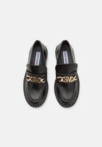 Mocassins en cuir noir à semelles épaisses avec un détail de chaîne dorée portant les initiales "SM" sur le dessus, dotés de semelles intérieures Steve Madden.