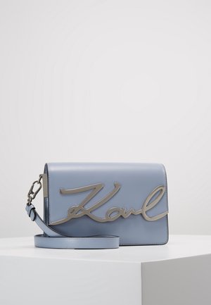 Sac bandoulière en cuir bleu clair avec la signature "Karl" en métal argenté sur le devant, sangle amovible, présenté sur une surface blanche.