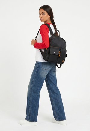 Kvinde med langt flet, iført rød og hvid langærmet skjorte, blå jeans, hvide sneakers, bærende på en sort rygsæk med forlommer.