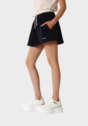 Donna che indossa una gonna sportiva nera con tasche, vita con coulisse bianca e sneakers bianche con suola alta, in piedi di lato con una mano in tasca.