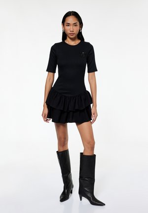 Vrouw die een zwarte mini-jurk met korte mouwen en ruches draagt, met een broche en zwarte kniehoge laarzen met hakken, staande op een witte achtergrond.