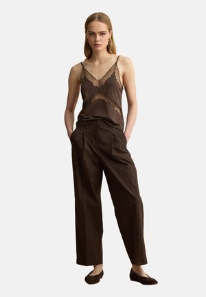 Woman wearing dark brown lace-trimmed camisole, loose brown trousers, and dark ballet flats, standing with hands in pockets against white background.