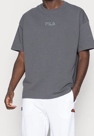 Homme portant un t-shirt gris FILA et un pantalon blanc avec un petit logo orange et noir sur la poche droite, debout avec une main dans la poche.