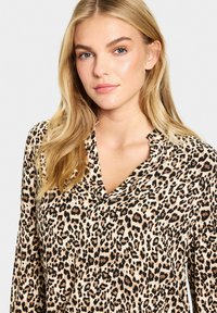Blouse à imprimé léopard en tissu doux et texturé, avec un décolleté en V profond et des manches longues. Les couleurs incluent des taches beige, noires et brunes.