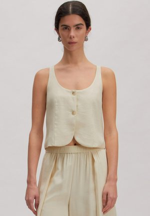 Frau trägt ärmelloses cremefarbenes, geknöpftes Oberteil und passende hochtaillierte, weite Hosen vor einem einfarbigen Hintergrund.