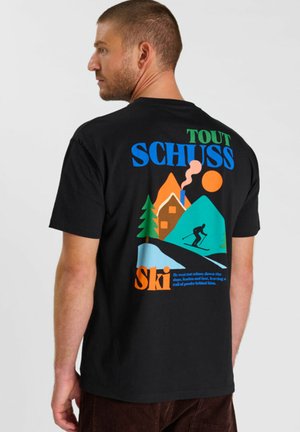 Uomo con t-shirt nera con grafica colorata a tema sci con montagne, sciatore, casa, albero, sole e testo "TOUT SCHUSS Ski" sul retro.