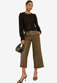 Top lungo a maniche nere con texture, abbinato a pantaloni a gamba larga in finta pelle verde oliva. Tessuto trasparente e forma aderente. Accessori includono una borsa nera.