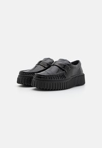 Zwarte loafers van glanzend leer met een textuurafwerking, voorzien van een dikke, geribbelde rubberen zool en klittenbandsluiting.