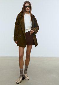 Brauner Corduroy-Mantel über einem weißen Pullover, kombiniert mit einem braunen Wildlederrock. Das Outfit umfasst beigemarmorierte Knöchelstiefel und graue Socken.