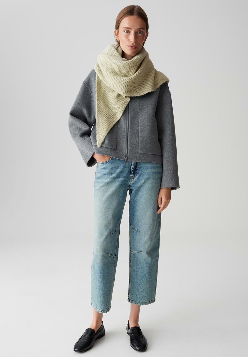 Femme portant une veste zippée grise, une écharpe en tricot crème, un jean bleu clair et des mocassins noirs, se tenant avec une main dans sa poche.