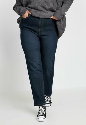 Jeans de pierna recta azul oscuro con una cintura alta, con detalles de costura dorada, combinados con zapatillas de lona en blanco y negro y un suéter de punto gris.