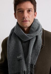 Écharpe grise à côtes en laine douce, présentant des bords frangés et une étiquette noire avec le logo "Marc O'Polo", enroulée autour du cou.