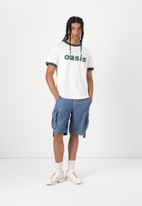 White t-shirt with green ringer collar and "oasis" text, paired with blue denim cargo shorts featuring side pockets. White sneakers complete the outfit.