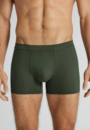 Męski tors w dopasowanych, ciemnozielonych bokserkach, stojący z ramionami swobodnie opuszczonymi na jasnym, jednolitym tle.