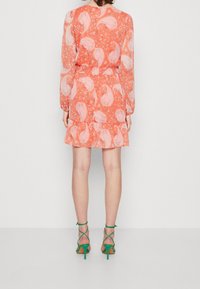 Korallenfarbens Kleid mit Paisleymuster, langen Ärmeln und Rüschensaum. Kombiniert mit grünen Riemchen-Heels, die einen offenen Rücken und eine schlanke Passform zeigen.