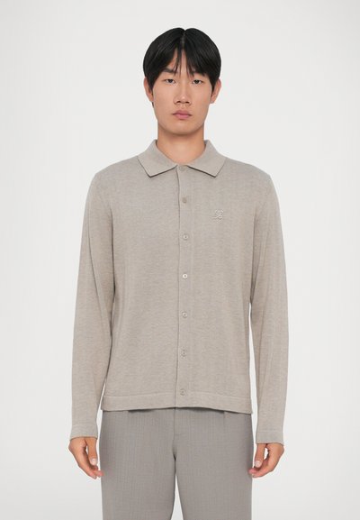 Polo a maniche lunghe beige realizzato in morbido tessuto a maglia, con colletto, chiusura a bottoni e un logo discreto sul lato sinistro.