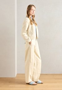 Krämfärgad, fluffig cardigan över en ljusblå topp, parat med lösa krämfärgade byxor och ljusblå sandaler med en brun sula.