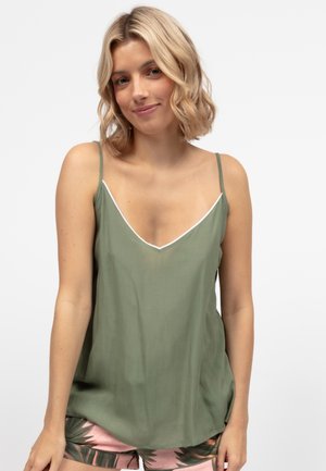 Blondynka w zielonym topie bez rękawów z białym obszyciem i różowych szortach w zielony wzór liści, uśmiechająca się na jednolitym tle.