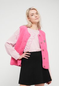 Ροζ χνουδωτό γιλέκο πάνω από ένα ανοιχτό ροζ πουλόβερ με προφίλ σχέδιο, σε συνδυασμό με μια μαύρη φουσκωτή φούστα. Διαθέτει χρυσά κουμπιά και υφασμάτινη υφή.