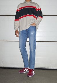Hombre de pie con la mano en el bolsillo, vistiendo una camiseta de rayas beige, roja y negra de manga larga, jeans rotos azules y zapatillas Adidas rojas.