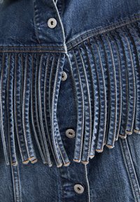 Gros plan d'une veste en denim montrant des franges verticales cousues sur le devant de la poitrine et des boutons argentés visibles le long de la couture centrale.