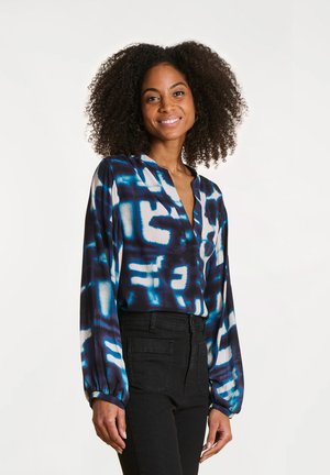 Blouse à manches longues avec un motif abstrait bleu et noir, dotée d'un col en V et d'une coupe ample, associée à un pantalon noir taille haute.