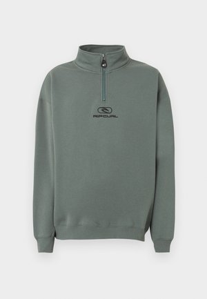 Jeune homme debout portant un sweat vert à demi-fermeture éclair avec le logo "Rip Curl" et un pantalon cargo noir sur un fond clair uni.