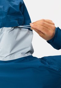 Giacca blu e grigia con una finitura testurizzata. Una mano regola il cordoncino elastico in vita, evidenziando un dettaglio di design funzionale.