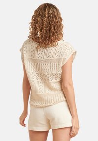 Gehaakte beige trui met korte mouwen, open haakpatronen en een relaxte fit, gecombineerd met crèmekleurige shorts, wat de textuur van het ontwerp accentueert.