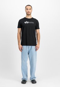 Sort kortærmet t-shirt med hvid "Alpha Industries" logo, parret med lysblå denimjeans og sorte slip-on sko.
