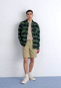 Grøn og sort ternet flannel skjorte, beige t-shirt, beige shorts og lyse højtop sneakers præsenteret mod en hvid baggrund.