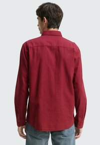 Chemise rouge à manches longues en tissu texturé, dotée d'un col à boutons et de détails subtils, associée à un pantalon en denim bleu.