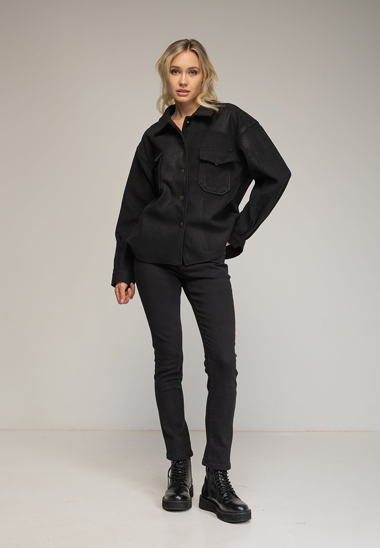 Camisa chaqueta negra de corte oversize con botones frontales y bolsillo en el pecho, combinada con jeans negros ajustados y botas negras con cordones. Textura suave.