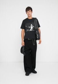 T-shirt gráfica preta apresentando um design de baterista, combinada com calças largas pretas. Os acessórios incluem um cinto preto e uma bolsa de ombro.