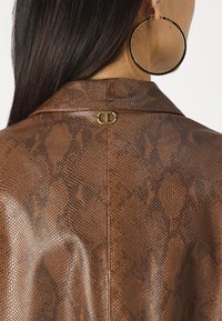 Barna, kígyó bőrére emlékeztető textúrájú dzseki, gallérral, arany színű fém díszítéssel a hátán. Nagy arany karika fülbevaló látható.