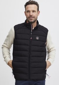 Svart polstret vest med ståkrage, glidelås foran, sidelommer og logodetalj. Bæres over en beige genser.