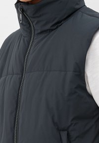 Schwarze ärmellose Steppweste mit hohem Kragen, Reißverschluss vorne und gestepptem Textil. Einfaches Design ohne sichtbare Hardware oder Muster.
