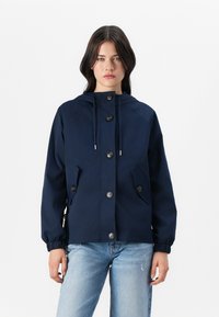 Marineblå jakke med hette, frontknapper og to sidelommer. Har elastiske mansjetter og et enkelt, lett design.