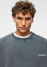 Grijze crewneck sweatshirt met een textuurafwerking en een witte "ESSENCE PROJECT" logo op de borst, gelaagd over een wit T-shirt.