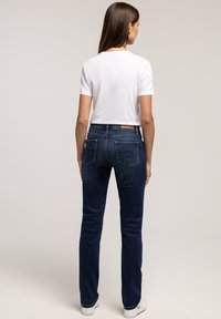 Kobieta stojąca plecami w prostym białym t-shircie, ciemnoniebieskich dżinsach o prostym kroju i białych sneakersach na jednolitym tle.